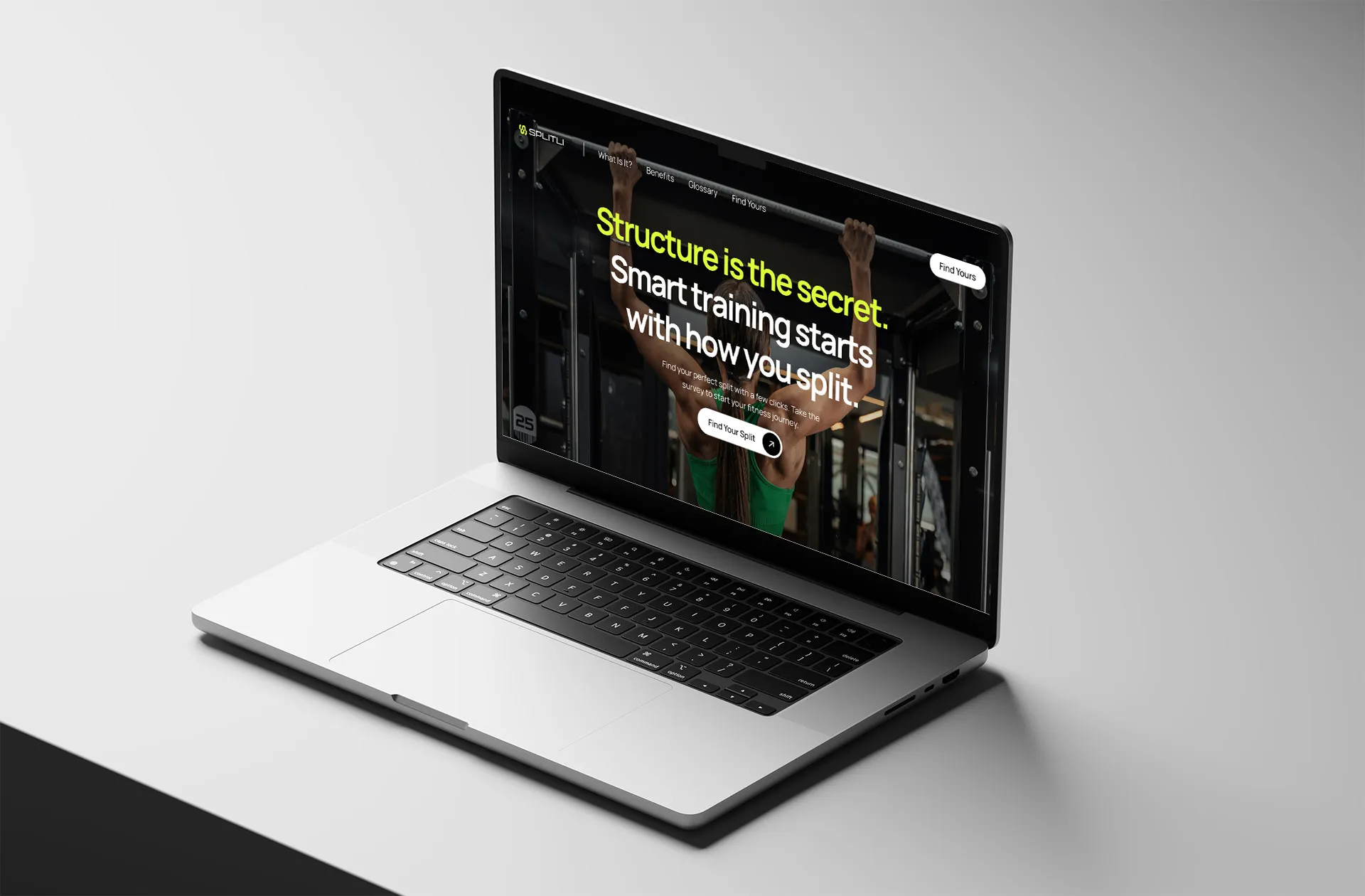 A silver Apple MacBook Pro 16-inch laptop is displayed with Splitli’s hero spread pictured on the screen. The laptop is on a white table surface, the scene is monochromatic aside from the laptop screen. The spread contains the heading “Structure is the secret. Smart training starts with how you split.” The opening image is of a woman, pictured backside in a green training bra performing a pull up. 