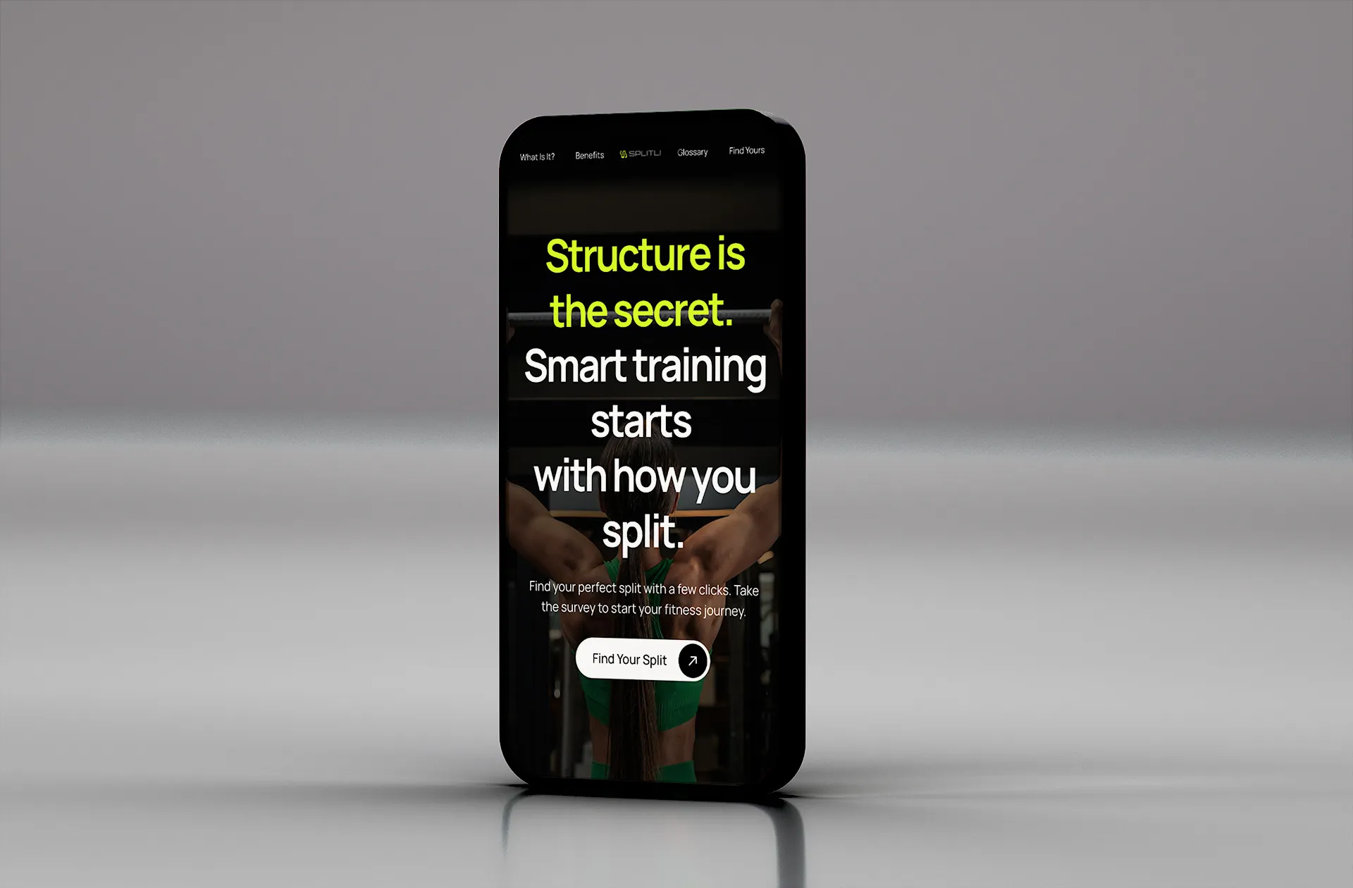 A silver iPhone 16 Pro is displayed with Splitli’s hero spread pictured on the screen. The phone is on a white table surface, the scene is monochromatic aside from the phone screen. The spread contains the heading “Structure is the secret. Smart training starts with how you split.” The opening image is of a woman, pictured backside in a green training bra performing a pull up. 